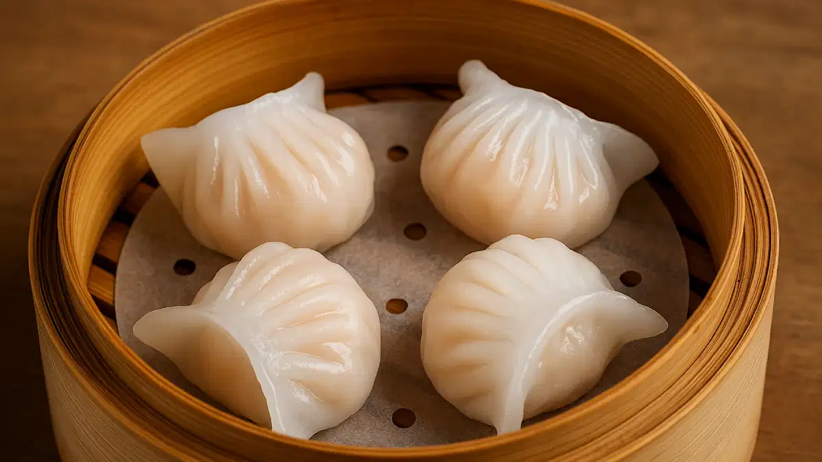 Nampan bambu berisi empat hakau dimsum berwarna putih susu, terletak di atas meja kayu dengan lapisan kertas berlubang di bawahnya.